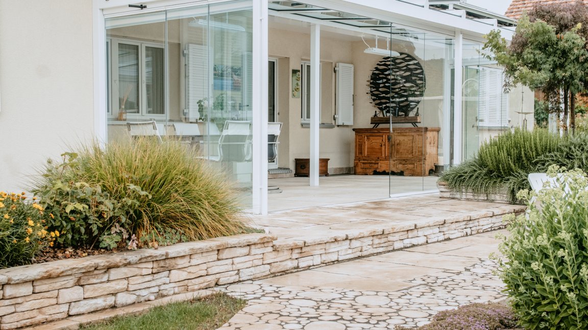 Jardin d’été blanc avec toit en verre et vitrage coulissant-pivotant COVER sur une maison de plain-pied, chemin en dalles rondes