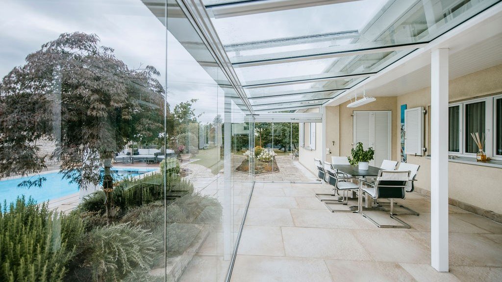 Jardin d’été avec toit en verre et vitrage coulissant-pivotant COVER, coin repas et vue sur la piscine