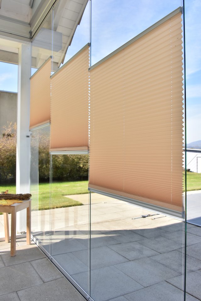 Beige PROTECT-Plissees an einer rahmenlosen Glasfront eines Sitzplatzes, teilweise heruntergelassen