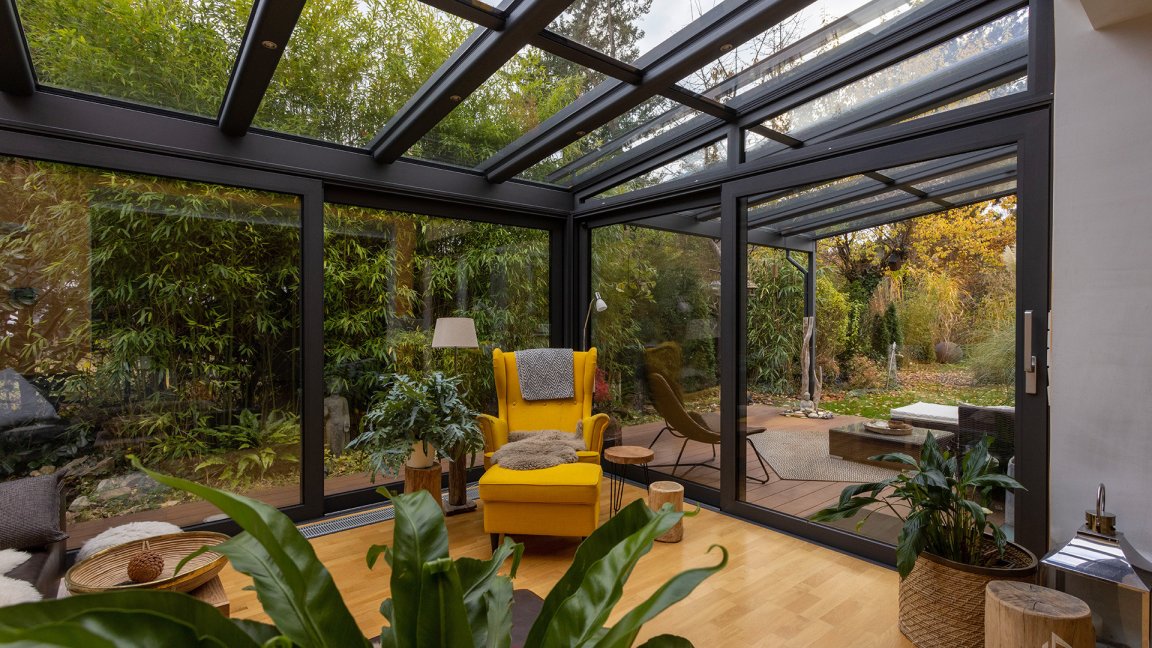 Jardin d’hiver RELAX chaleureux avec toit en verre, fauteuil jaune et parquet, vue sur un jardin d’automne