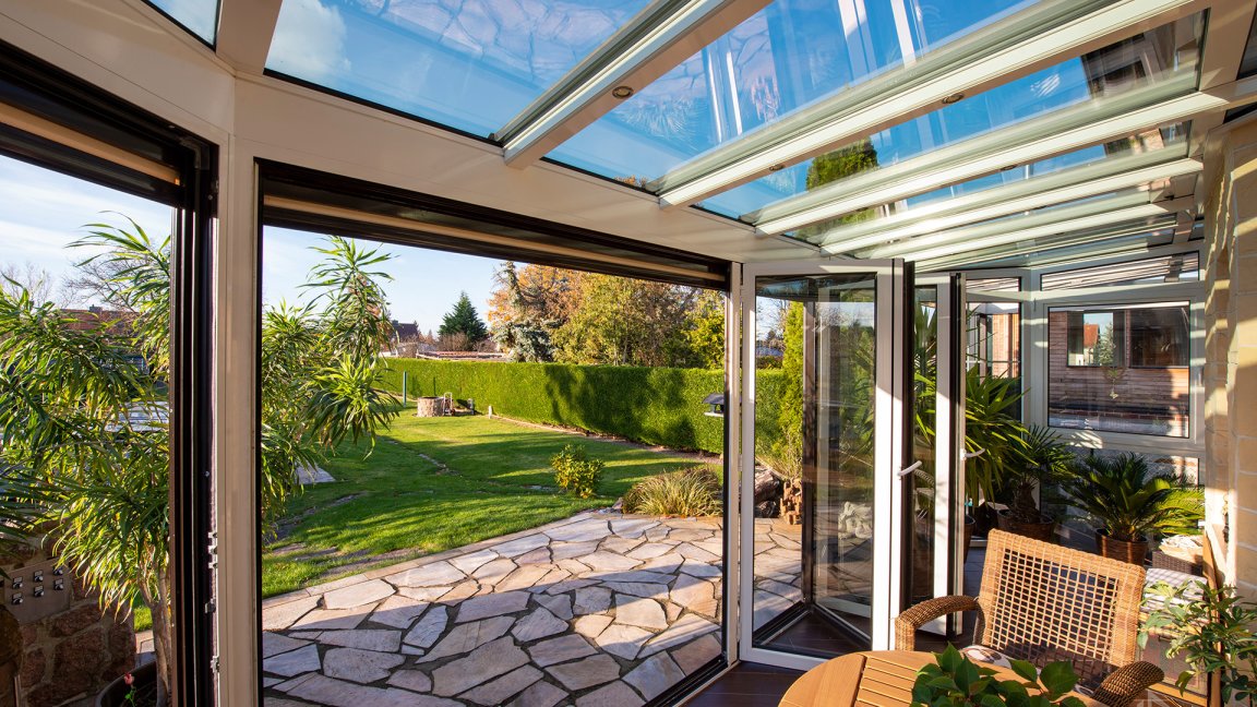 Vue depuis un jardin d’hiver RELAX blanc avec toit en verre, par la porte ouverte sur le jardin