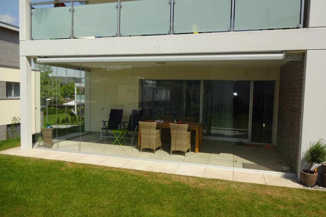 Vitrage de terrasse sans cadre au rez-de-chaussée d’un immeuble, sous un balcon à garde-corps en verre