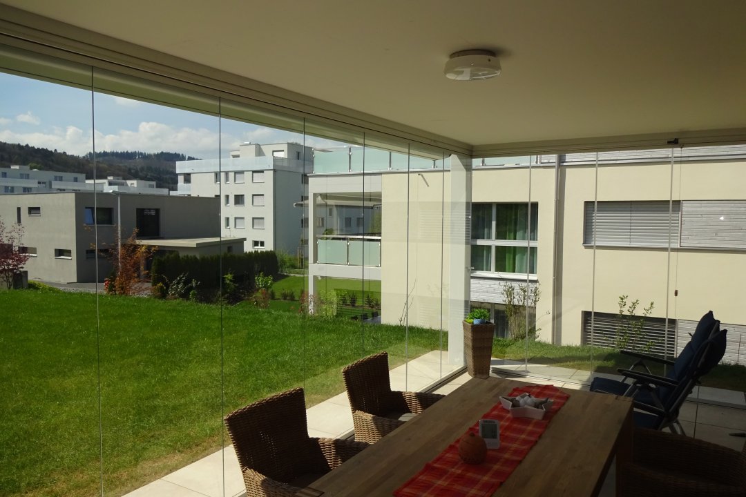 Vue à travers un vitrage de terrasse sans cadre, avec table à manger, sur la pelouse et les immeubles voisins, réalisation Fremet AG