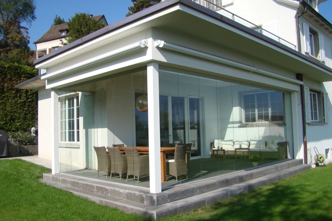 Terrasse couverte d’une villa fermée par un vitrage sans cadre, avec table et chaises en rotin, réalisation de Ledas GmbH