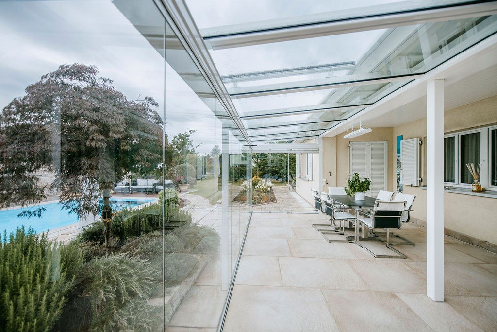 Toit en verre et vitrage COVER sans cadre protégeant la terrasse du vent et des intempéries, à côté d’une piscine