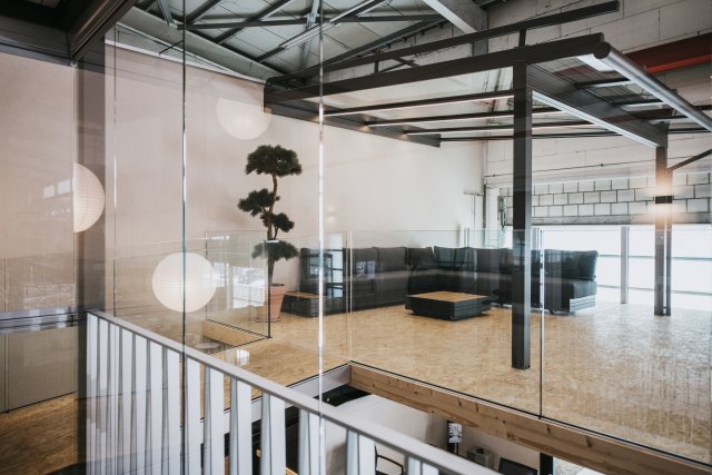 Vista oltre una ringhiera bianca, attraverso una parete in vetro, su un lounge in stile loft con divano scuro e travi in acciaio