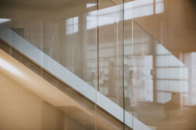 Scala con parapetto in vetro senza telaio in un ambiente interno luminoso, le superfici in vetro riflettono la facciata finestrata