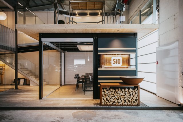 Cubo espositivo vetrato con logo SIO, tavolo da pranzo e braciere su legna da ardere sotto un soppalco nello showroom