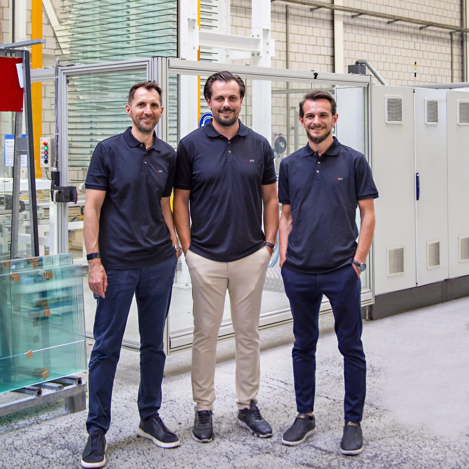 Drei Männer stehen in einer Produktionshalle zwischen Lagerwagen mit Aluminiumprofilen